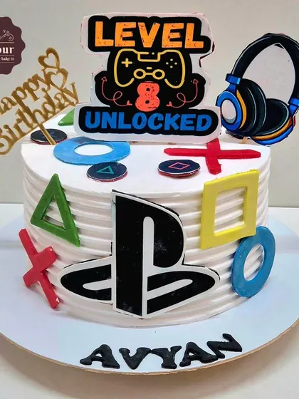 A PlayStation-themed cake with "Level 8 Unlocked" for a young gamer. The cake is decorated with the iconic controller button symbols and a headset topper.