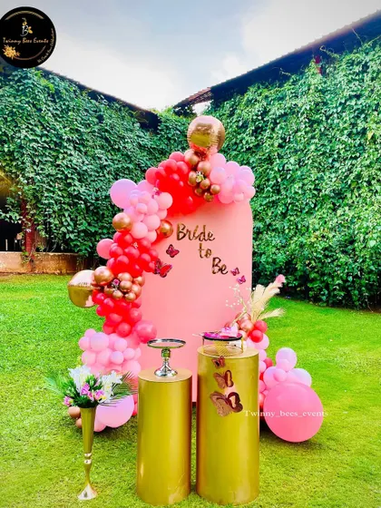A "Bride to be" celebration in a beautiful garden setting. The decor features a pink arched backdrop with a vibrant red and pink balloon garland and elegant gold pedestals.