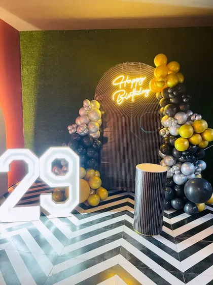 A modern setup for a 29th birthday. The decor includes a black, gold, and silver balloon garland against a unique backdrop and chevron-patterned floor.