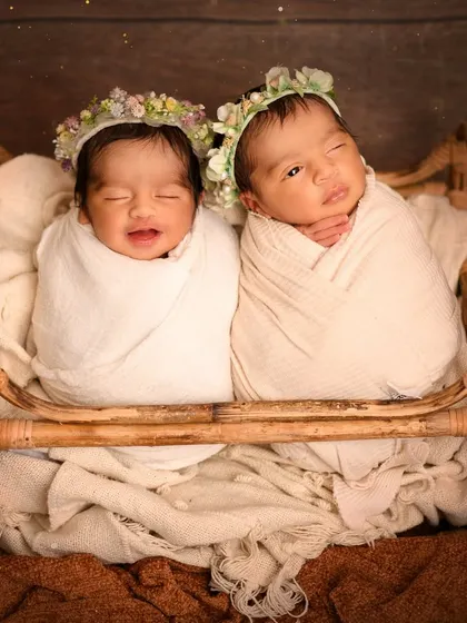 Two newborn twins, swaddled in white and wearing floral wreaths, are pictured sleeping together in a rustic wooden crib. This heartwarming shot captures the special bond between twins from their very first days.