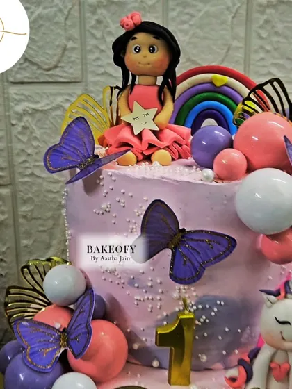A close-up of the two-tier unicorn cake, showing the handmade fondant doll, purple butterflies, and colorful orbs. Every detail is crafted with love for the little one's special day.