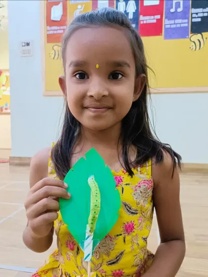 A young girl in a bright yellow dress holds her green leaf and caterpillar craft. Our activities often tie into themes about nature and the world around us.