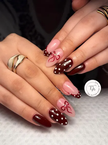 A chic and stylish manicure featuring a deep maroon polish, white polka dots, and delicate, hand-painted bows on a nude base.