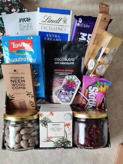 A delightful Diwali hamper filled with a mix of treats and self-care items. It includes assorted chocolates, wafers, hot chocolate mix, a scented candle, a neem wood comb, and more.