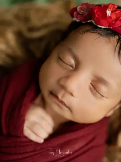A serene close-up of a sleeping baby girl, wrapped in a rich scarlet fabric with a matching floral headband. The focus is entirely on her angelic features.