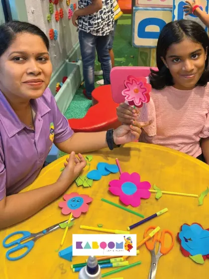 A girl proudly holds up the pink paper flower she just finished making, while our staff member looks on with a smile. We love seeing their creative confidence grow.