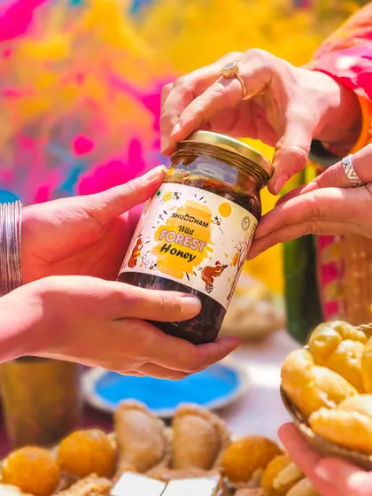 A shot focusing on the act of gifting, with hands exchanging a jar of honey against a colorful Holi background.