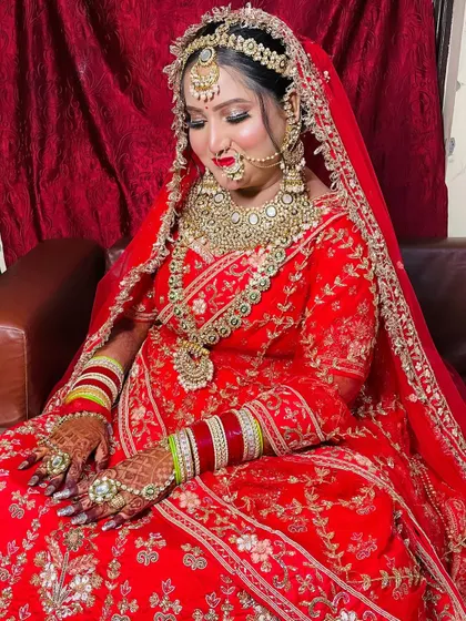A full view of the bride seated, looking like royalty in her red lehenga with intricate gold work and layered jewelry.