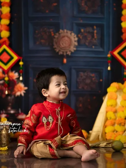The joy of celebration shines in this little boy's smile. Dressed in a festive red kurta and dhoti, he is surrounded by a traditional setup of marigold garlands and brass lamps.