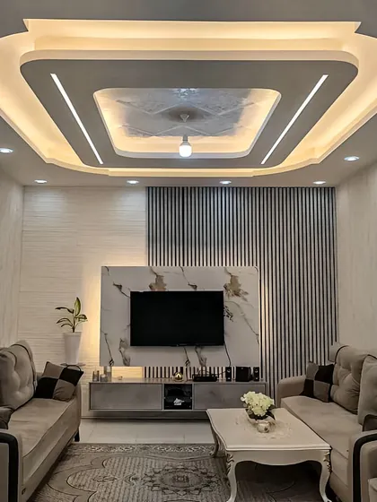 This living room setup features a compact TV unit against a wall with mixed textures of fluted panels and white marble. The design is complemented by an elaborate false ceiling with integrated lighting.