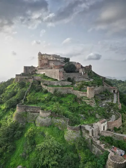 A dramatic, wide-angle shot of Kumbhalgarh Fort under a cloudy sky. This perspective showcases the fort's dominance over the surrounding Aravalli hills and captures a sense of its timeless, enduring presence.