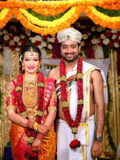 A classic portrait of the bride and groom on their wedding day. They are dressed in beautiful traditional South Indian wedding attire, standing before a colorful floral backdrop.