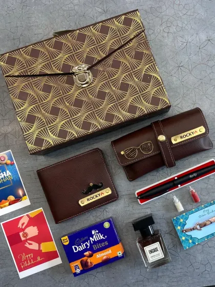 A premium Rakhi gift box with a patterned lid. Inside, it holds a personalized wallet and glasses case, a pen, perfume, chocolate, and rakhi cards.