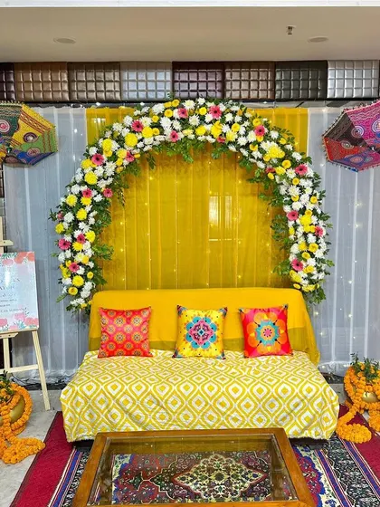 A cozy Haldi setup with a sofa decorated with colorful printed cushions. The backdrop is a floral arch against a yellow drape, with Rajasthani umbrellas adding a festive touch.
