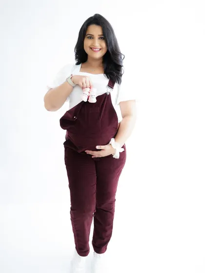 A fun and casual announcement. The mom-to-be wears cute overalls and holds up a tiny pair of baby shoes, smiling brightly.