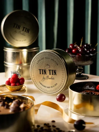 The signature "Cheesecake in a Tin" from Bastian. This shot showcases the elegant branding and packaging, surrounded by fresh berries to hint at the flavours inside.