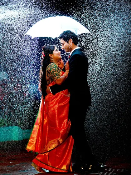 A romantic pre-wedding style shot in the rain under an umbrella. The bride's red saree and the groom's suit create a classic, cinematic look.