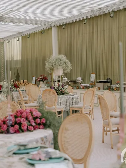 A wide view of the luncheon space, showing the striped marquee ceiling and sage green curtained walls. The layout provides a mix of round tables for an intimate and communal dining experience.