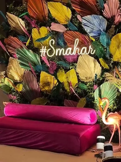 A modern and artistic Mehendi seating area. The backdrop is a burst of colourful painted palm leaves with a custom neon hashtag, paired with a plush magenta sofa and flamingo accents.