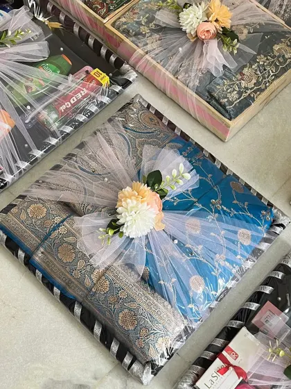 A close-up of a beautifully wrapped trousseau gift. A rich blue banarasi silk saree is packed in a striped tray, finished with a delicate tulle bow and a floral accent.