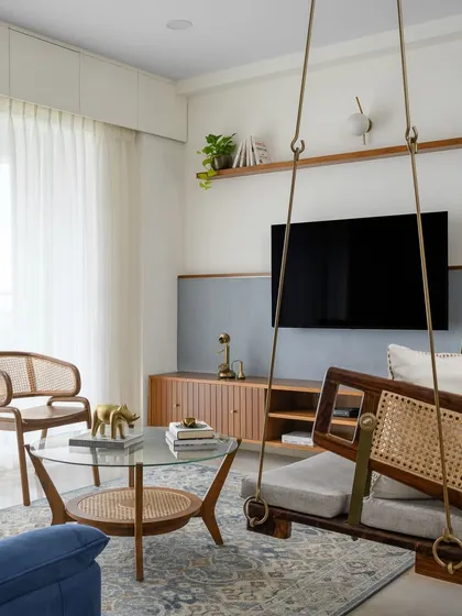 The 'Malhar' living room features a sleek veneered TV unit against a textured half-wall. The wood and cane swing and chairs complete the ensemble, creating a harmonious blend of modern and traditional elements.