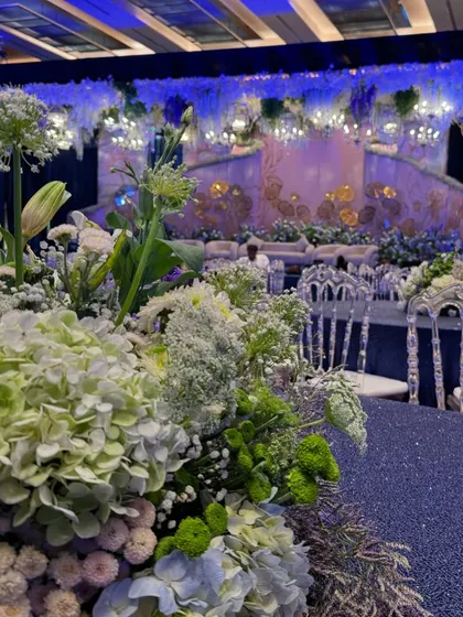 A close-up of a table centerpiece, featuring hydrangeas and other white and green florals, set against the backdrop of the blue-lit reception stage. This shows how the floral design complements the overall color scheme.
