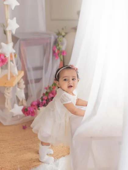 A baby girl pulling herself up to stand, framed by soft white curtains and bougainvillea. Milestone sessions are all about capturing these exciting new skills.