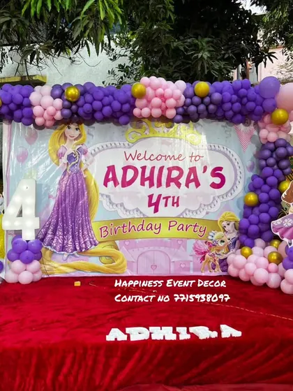 A Rapunzel-themed 4th birthday party setup. The decor includes a large backdrop featuring the princess, a purple and pink balloon arrangement, and a light-up number '4'.