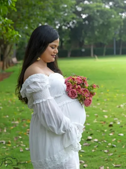 A stunning solo portrait of the mama-to-be in a white off-shoulder dress, holding a bouquet of pink roses. She looks serene and beautiful.