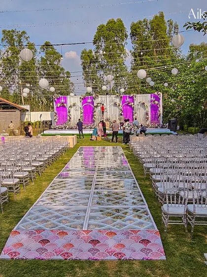 A wide view of an outdoor wedding ceremony setup, featuring a long, transparent aisle filled with flowers. The stage is decorated with purple and white drapes and floral arrangements, with paper lanterns hanging overhead.