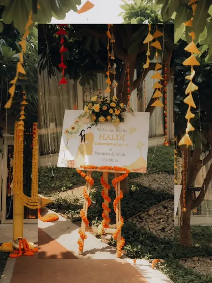 A personalized welcome sign for Purnima and Rohit's Haldi, set amidst trees decorated with marigold garlands.