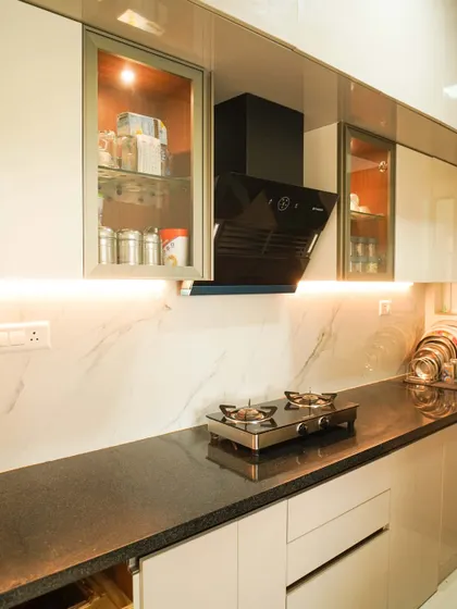 This kitchen combines glossy beige cabinets with a white marble-effect backsplash and countertop. The glass-fronted upper cabinets with internal lighting provide an elegant display area, while the under-cabinet lighting ensures a well-lit workspace.