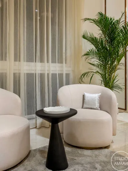 This cozy corner shows our focus on creating comfortable, stylish nooks. Two plush, curved armchairs in a soft neutral fabric are paired with a sculptural black side table. A large potted palm and sheer curtains add to the light and airy feel.