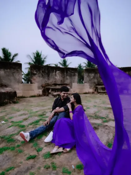 A playful and romantic moment captured during a pre-wedding shoot. The flowing purple saree adds a vibrant splash of color and movement to the scene.