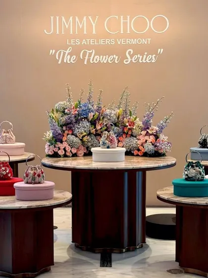 A multi-level product display showcasing Jimmy Choo's new collection. Each handbag is presented on its own table, surrounding a central, grand floral arrangement that unifies the entire presentation.