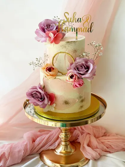 A romantic two-tier cake with a soft pink watercolor effect and gold leaf details. This engagement cake is adorned with purple and peach roses, baby's breath, and a gold hoop accent.