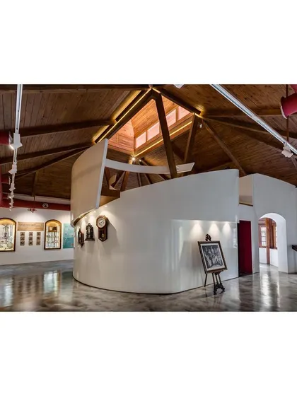 The central armature of the museum, a curved white volume containing an AV room and a staircase leading to a mezzanine. This element organizes the visitor's path and sits in contrast to the exposed wooden roof structure of the original building.