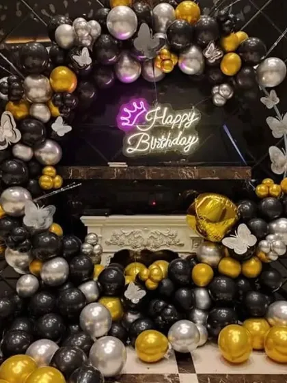 A birthday party with a black, gold, and silver balloon arch. The decoration includes a "Happy Birthday" neon sign with a crown and butterfly accents.