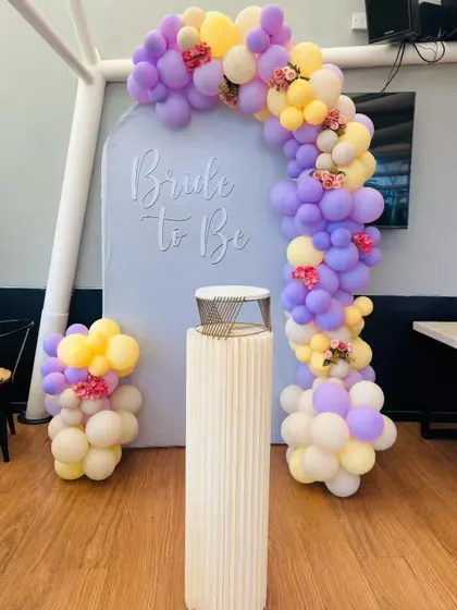 This pastel-themed "Bride to Be" decor is both elegant and sweet. The custom-arched backdrop is decorated with a beautiful balloon garland in shades of purple, yellow, and cream, with small floral accents.