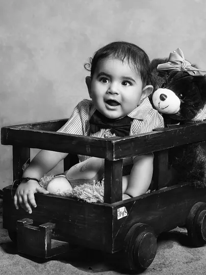 A baby boy in a wooden cart with his teddy bear. This black and white photo has a classic, nostalgic feel.