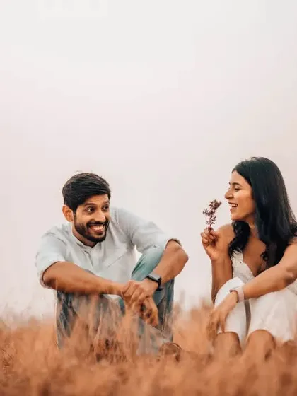 A sweet and simple pre-wedding moment in a field, where the bride-to-be playfully offers her partner a flower.