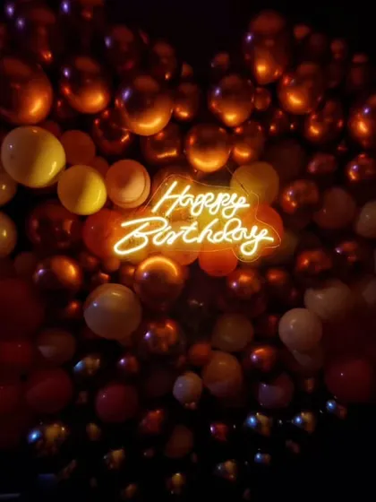 A close-up of the 'Happy Birthday' neon sign nestled within a heart-shaped balloon arrangement. The warm orange glow creates a cozy and inviting atmosphere for the celebration.