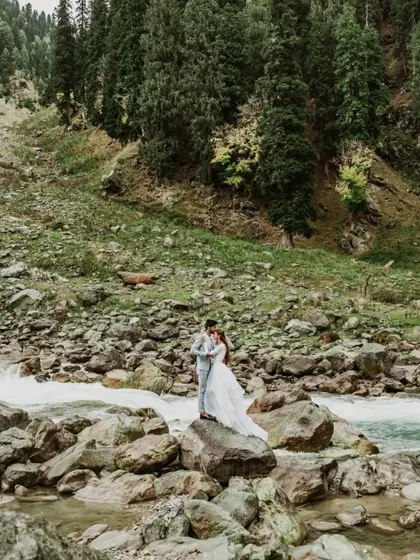 A wide shot capturing the grand scale of the Kashmir landscape as a couple stands by a river. This image highlights the epic, adventurous feel of a destination pre-wedding shoot.