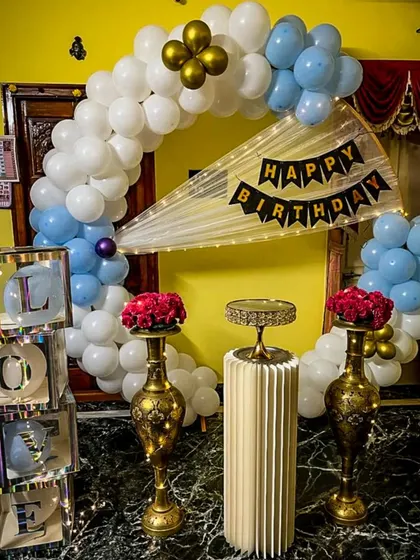 We create loving tributes for your special occasions. This home setup features a blue and white balloon arch, elegant brass vases with red roses, and illuminated "LOVE" display boxes for a heartfelt anniversary or birthday celebration.