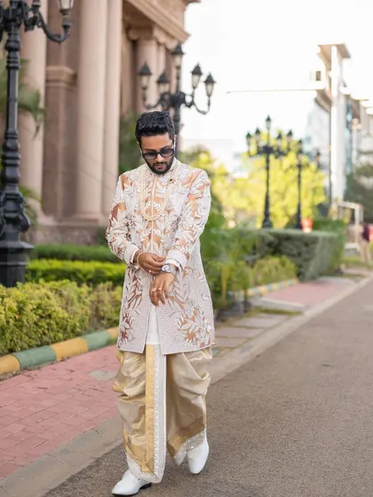 This groom is pictured in a custom off-white sherwani with elaborate gold and white embroidery. The outfit is paired with a silk dhoti, creating a classic and sophisticated look suitable for a wedding ceremony.