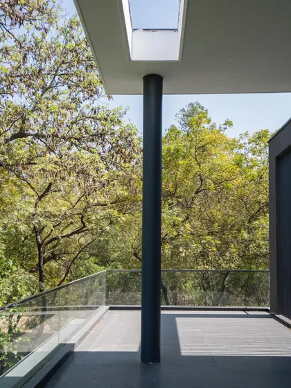 A balcony at Triveni Farmhouse offers an immersive experience of the surrounding tree canopy. A simple black column provides structural support while a cutout in the roof above allows sunlight to filter through, connecting the space to the sky.