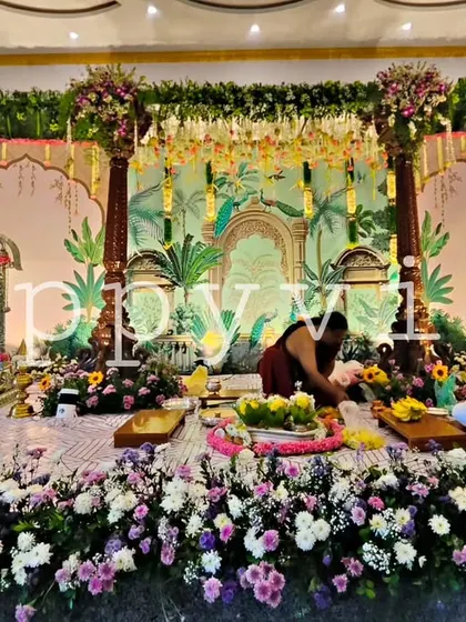 A wide view of a traditional naming ceremony in progress. The decor features a hand-painted backdrop with tropical motifs and an abundance of fresh flowers on the floor, creating a sacred space.