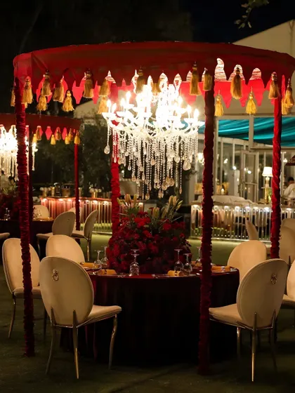 An elegant outdoor dining setup for a formal ceremony. Each table features a miniature canopy with red tassels and a crystal chandelier, creating individual, intimate dining spaces.