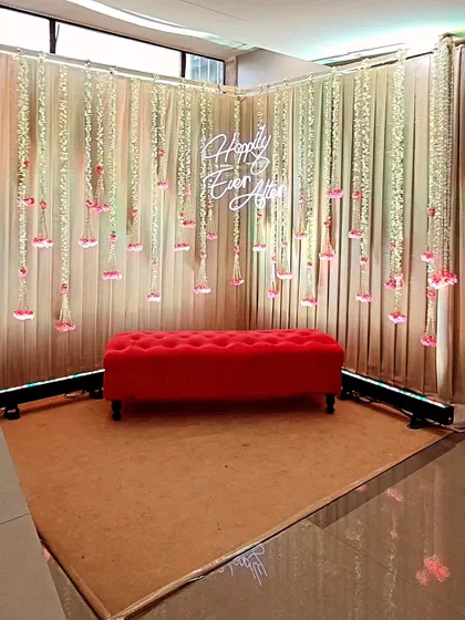 A perfect selfie corner with a "Happily Ever After" neon sign. The backdrop of hanging floral strings and a bright red sofa creates a romantic and fun photo opportunity for the couple and guests.