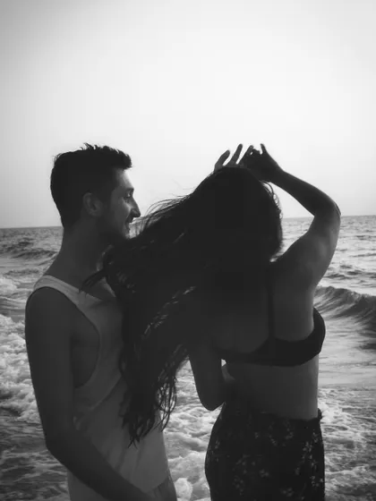 A candid black and white photograph capturing a gentle, unposed interaction by the sea. I look for these small gestures and glances that tell the real story of a couple's connection.